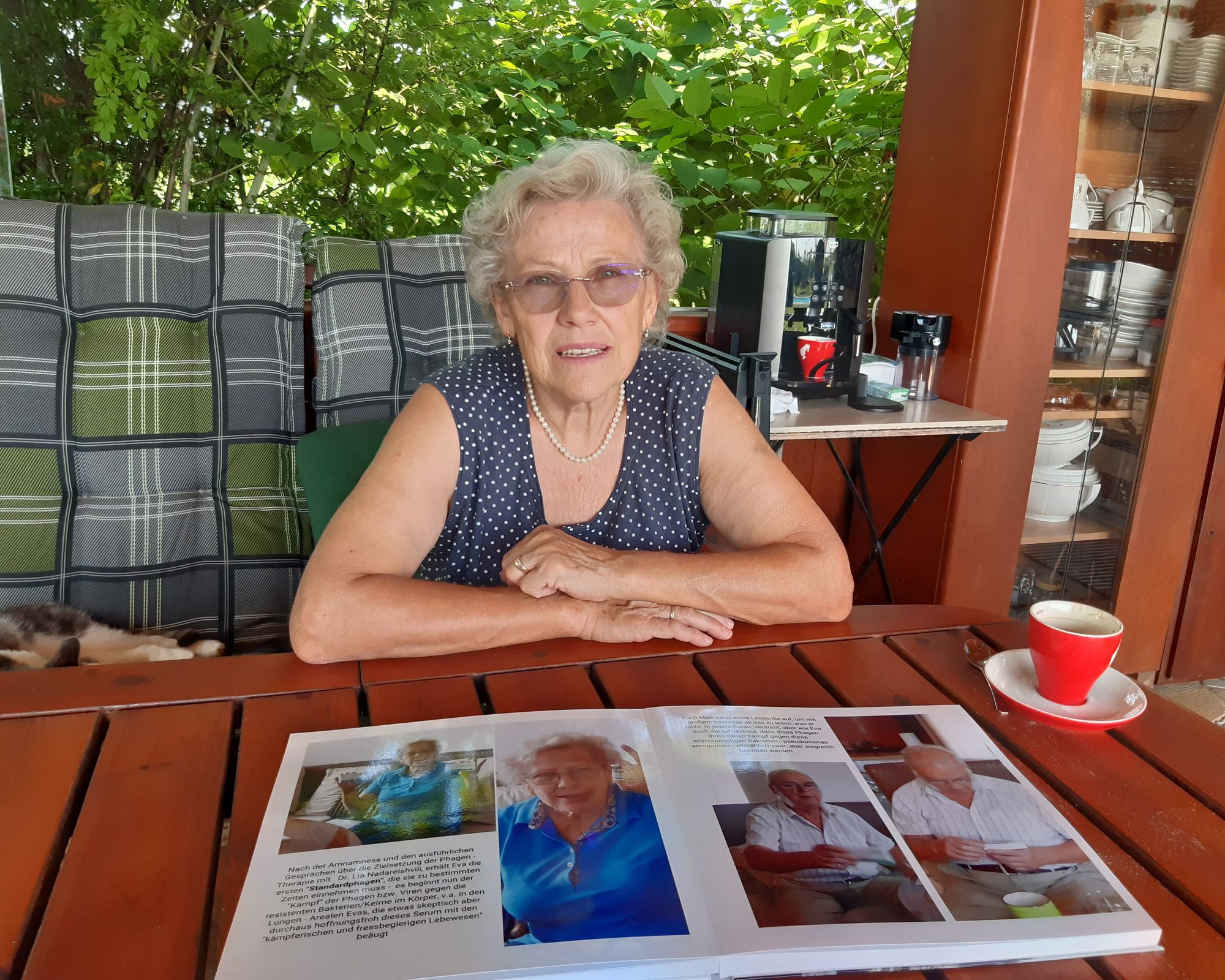 Eine ältere Frau sitzt auf einer gepolsterten Bank vor einer Hecke. Auf dem Holztisch vor ihr liegt ein großes Buch. Die aufgeschlagene Seite zeigt unter anderem zwei Fotos derselben Frau, wie sie eine Medikamentenpackung bzw. eine kleine Ampulle in die Kamera hält.