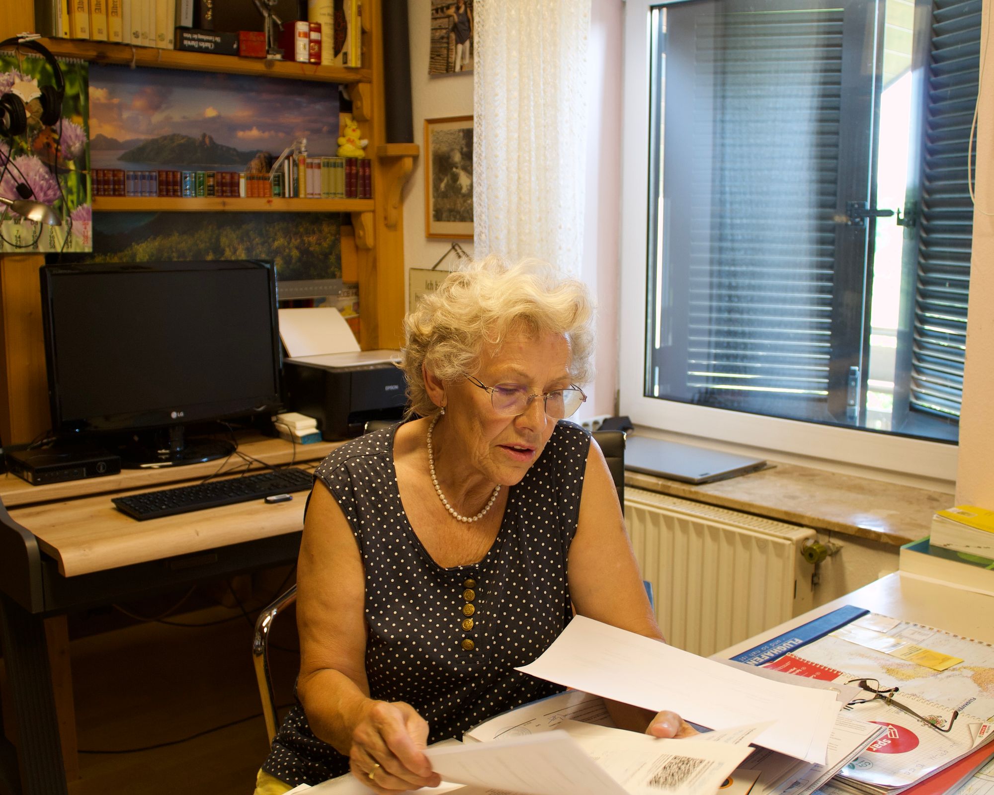 Eine ältere Frau in einem getupften Shirt sitzt an einem Schreibtisch und blättert durch bedruckte Papiere. Im Hintergrund sind ein Bücherregal, ein Computer, ein Drucker und ein Fenster mit geschlossenen Rollos zu sehen.