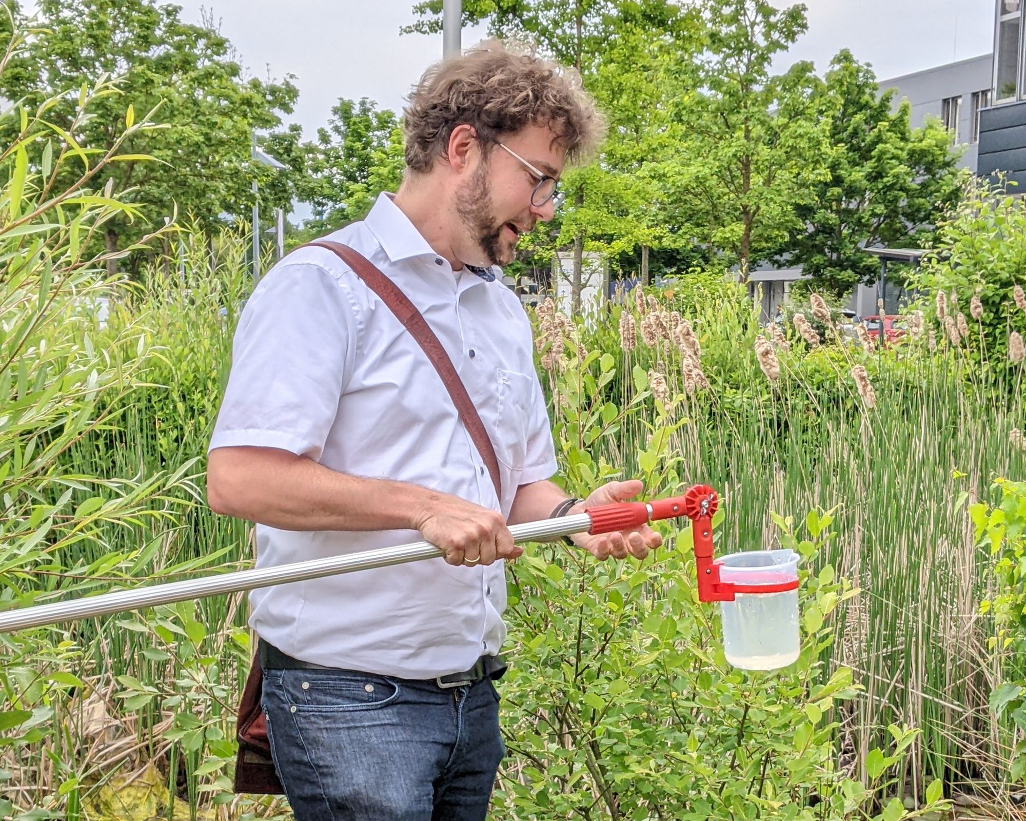 Ein Mann in Kurzarm-Hemd und Jeans steht zwischen hohem Schilf und Büschen. Er hält eine Teleskopstange, an der ein durchsichtiger Plastikbecher befestigt ist.